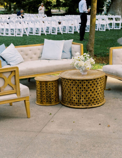 Outdoor seating area with wooden chairs, a loveseat, a round table, and white cushions; white chairs arranged in rows are visible in the background on grass.