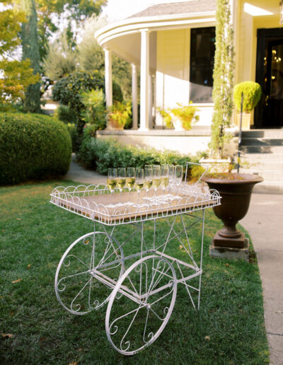 A white metal cart with wheels holds several empty wine glasses and a bowl, set on a lawn near a yellow house with a porch and greenery in the background.