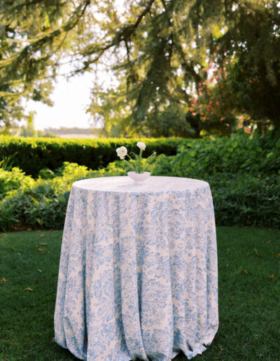 A small round table with a blue and white patterned tablecloth stands outdoors on grass, holding a small vase with white flowers and greenery. Lush trees and bushes are in the background.