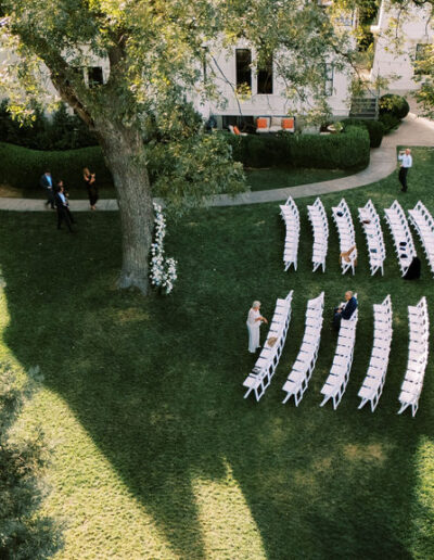 Aerial view of an outdoor wedding ceremony setup with white chairs arranged in rows on a lawn, a few people standing and sitting, and trees casting long shadows.