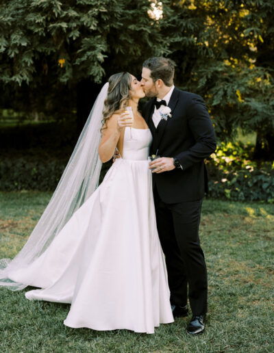 A bride in a white gown and veil kisses a groom in a black suit and bow tie outdoors, both holding drinks, with trees in the background.