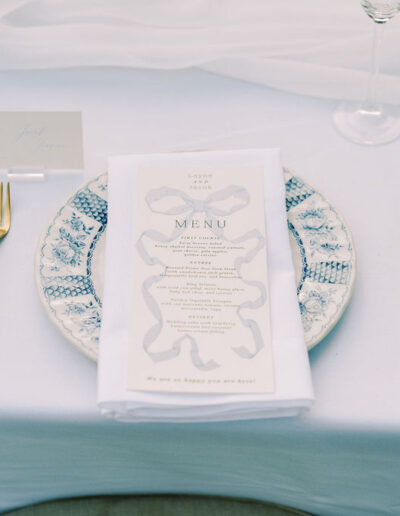 A place setting with a blue and white plate, gold cutlery, a menu on a white napkin, two wine glasses, and a name card on a white tablecloth.