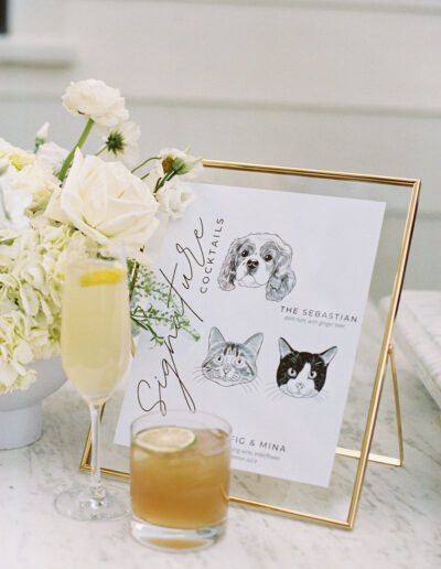 A framed cocktail menu with pet illustrations stands on a marble surface beside a white flower arrangement and two cocktails, one in a flute and one in a rocks glass.