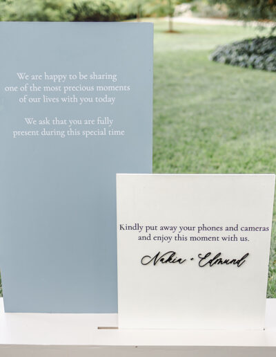 Two wedding signs on display outdoors; one requests guests to be present, and the other asks them to put away phones and cameras and enjoy the moment.