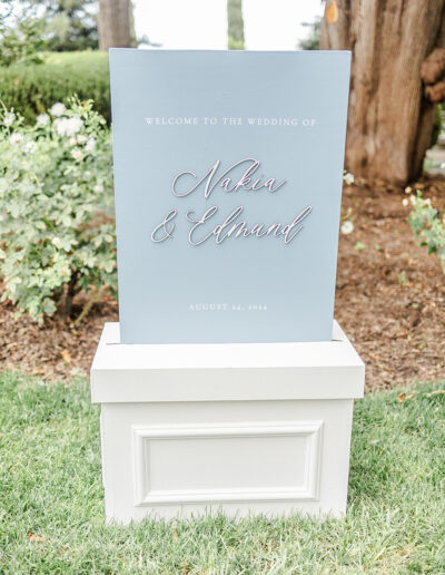A wedding welcome sign reads "Welcome to the wedding of Nakia & Edmund, August 24, 2024," displayed outdoors on a white stand.
