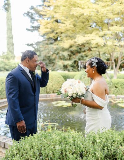 A bride in a white dress holding a bouquet smiles at a groom in a navy suit wiping his eye, standing by a small pond in a garden.