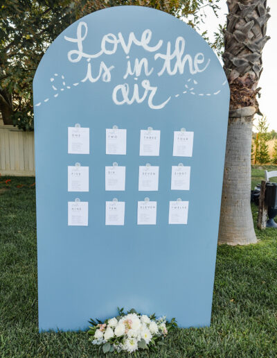 A blue sign with "Love is in the air" written at the top, displaying twelve clipped cards in rows, standing on grass with a white floral arrangement at the base.
