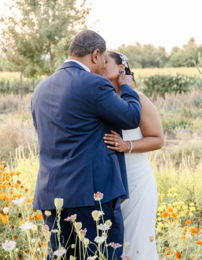A couple dressed in formal attire shares a kiss in a garden with orange and white flowers, with greenery and trees in the background.