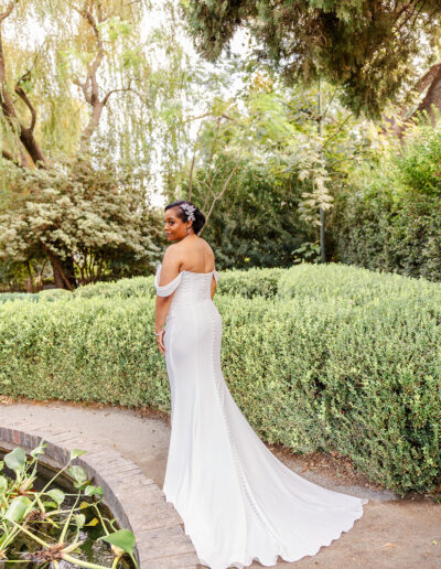 A woman in a white off-shoulder wedding dress stands on a garden path near a pond, surrounded by lush greenery and trees.