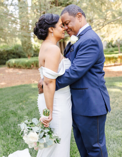 A bride and groom in formal attire embrace outdoors, touching foreheads and smiling, with the bride holding a bouquet of white roses and greenery.