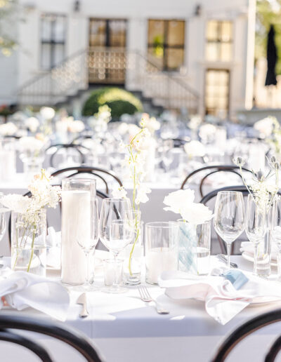 Long tables set for an outdoor event with white tablecloths, glassware, candles, and simple white floral centerpieces; a large building and greenery in the background.