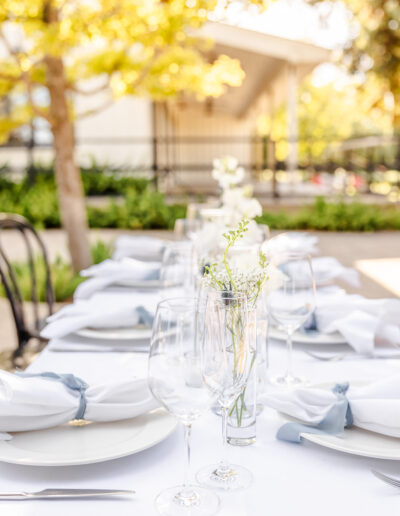 A white tablecloth dining table is set outdoors with plates, napkins, wine glasses, and small vases of white flowers, surrounded by greenery and soft sunlight.