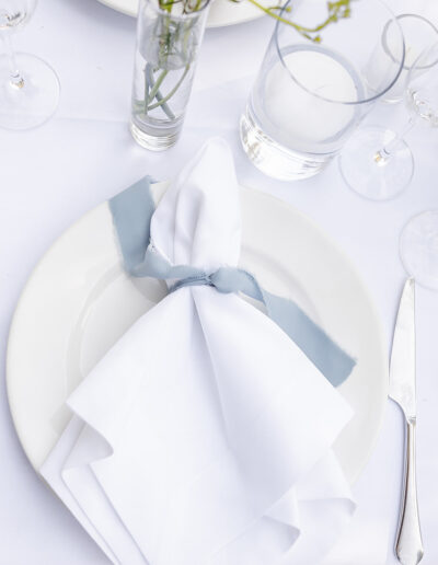 A white plate with a neatly folded white napkin and blue ribbon is set on a white tablecloth, surrounded by silver cutlery, glassware, and a small vase with flowers.