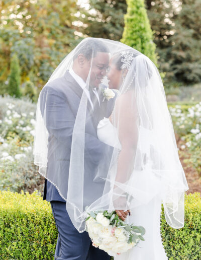 A bride and groom stand in a garden, facing each other and smiling under a shared veil. The bride holds a bouquet of white flowers.