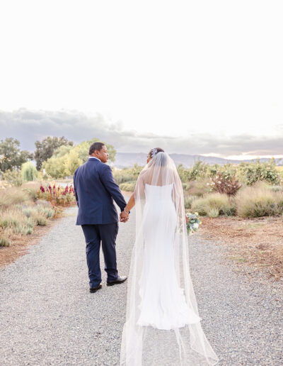 A bride in a white dress and veil holds hands with a groom in a navy suit as they walk down a gravel path in a garden setting.