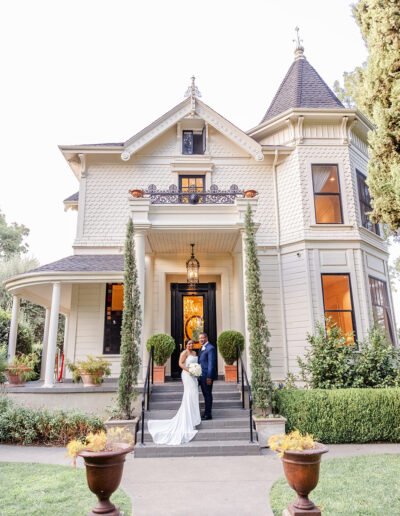 A couple in wedding attire stands on the front steps of a large, light-colored Victorian-style house surrounded by greenery.