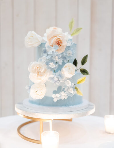 A light blue tiered cake decorated with white and peach sugar flowers and green leaves sits on a marble stand, surrounded by small lit candles.