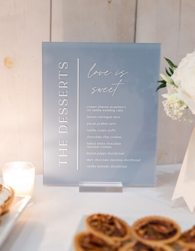 A dessert menu sign lists various sweets such as cream cheese strawberry, lemon meringue tarts, and chocolate chip cookies on a table with candles, flowers, and pecan tarts.