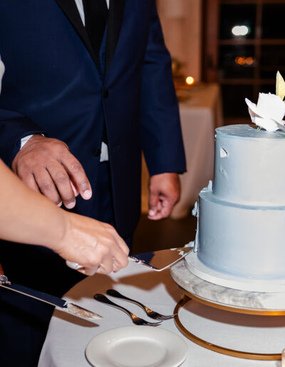 Two people, dressed formally, are cutting a two-tiered light blue cake with white floral decorations at an event.