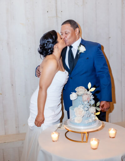 A couple dressed in wedding attire kisses beside a decorated cake on a table with lit candles, in front of a white wooden wall.
