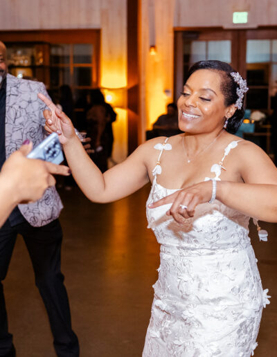 A woman in a white dress dances and smiles at an indoor event, with other people visible in the background.