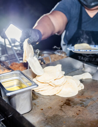 A person wearing gloves and a mask serves tortillas from a grill at a food stand, with various food trays and a plate visible.