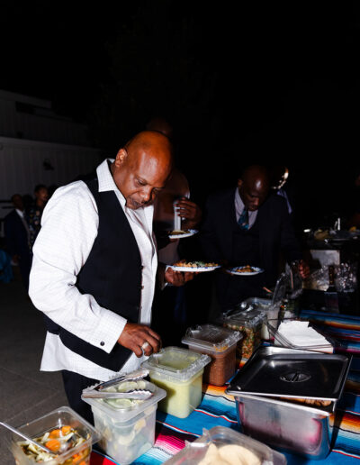 A man in a white shirt and black vest serves himself food from a buffet table outdoors at night.