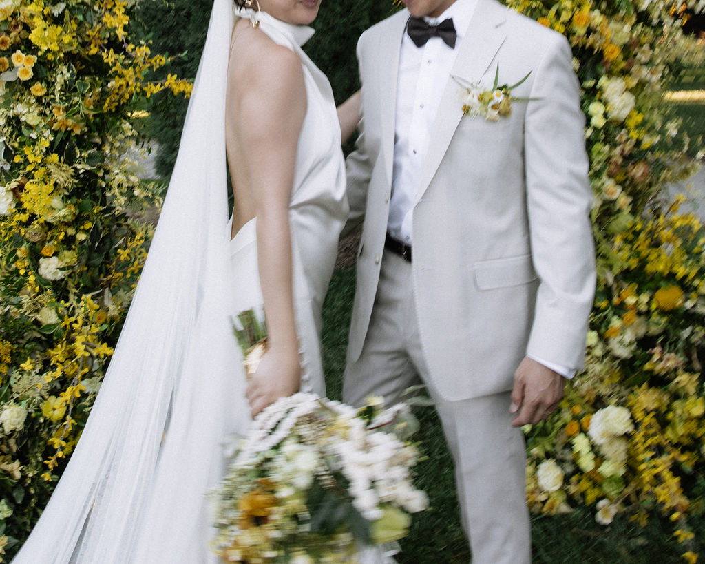 A bride and groom stand together outdoors in front of a floral arch with yellow flowers; the bride holds a bouquet and wears a veil, and the groom is in a light suit with a bow tie.
