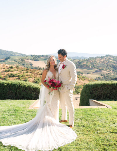 A bride and groom in formal attire stand together on a lawn, with green hills and clear skies in the background. The bride holds a bouquet of red flowers.