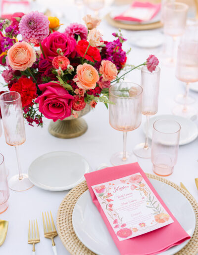 A table setting with a floral centerpiece of pink and orange flowers, pink glassware, gold cutlery, and a menu placed on a white plate with a woven charger.