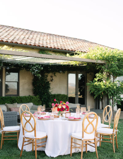 A round outdoor dining table with a white tablecloth, wooden chairs, and a floral centerpiece is set on a lawn near a patio and a vine-covered house.