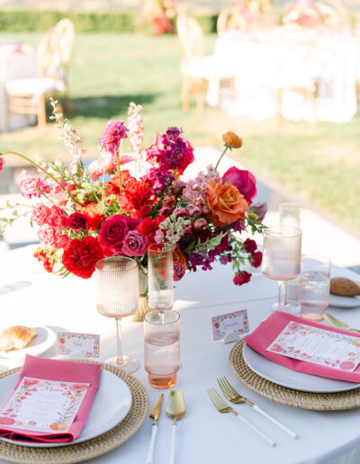A round table set for two with gold utensils, pink napkins, menus, place cards, and a vibrant floral centerpiece in red, pink, and orange hues.