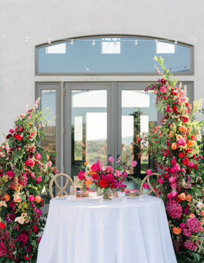 A round table with a white tablecloth is set for two in front of glass doors, surrounded by vibrant, colorful floral arrangements.