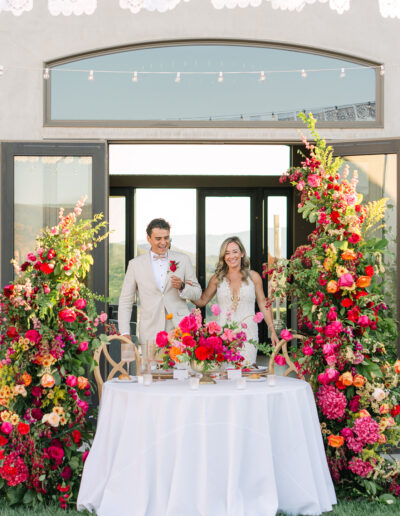 A bride and groom stand smiling behind a table with a white tablecloth, surrounded by vibrant red, pink, and orange floral arrangements in an outdoor setting.