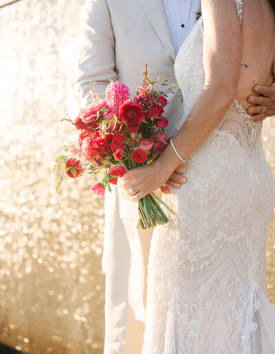A bride in a lace dress holds a bouquet of red and pink flowers, standing next to a groom in a light suit in front of a golden textured background.