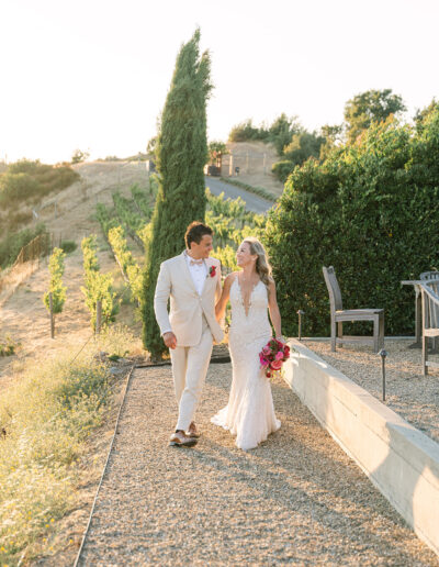 A bride and groom walk outside along a gravel path at a vineyard, holding hands and smiling at each other in the sunlight.