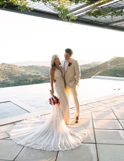 A bride and groom stand together by an outdoor pool, with hills in the background. The bride wears a long, fitted gown and holds a bouquet; the groom wears a light suit with a red boutonniere.