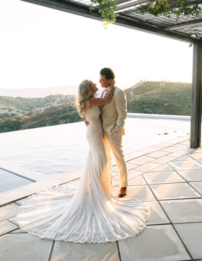 A bride and groom stand embracing by an infinity pool on a tiled terrace overlooking hilly countryside at sunset.