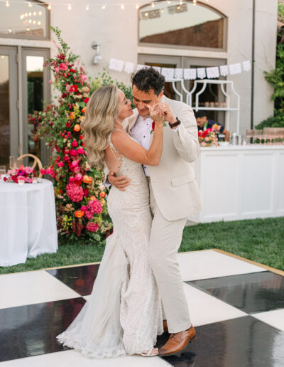 A bride and groom dance together on a black and white checkered floor at an outdoor wedding reception, surrounded by colorful floral decorations.