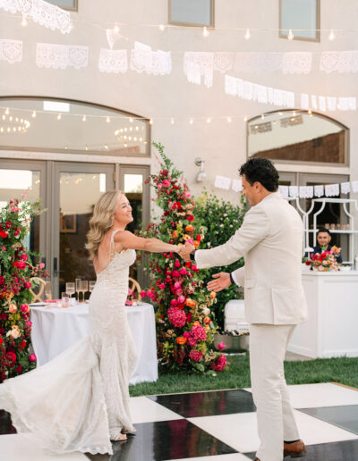 A bride and groom dance together on a black and white checkered floor at an outdoor wedding, with colorful flowers and string lights in the background.