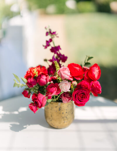 A gold vase filled with red and pink roses and other flowers sits on a white table outdoors, with greenery blurred in the background.