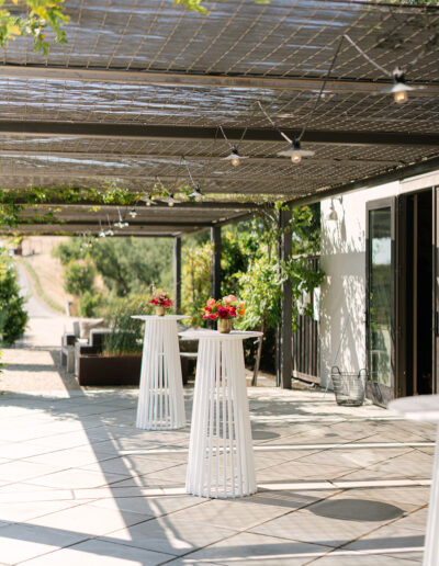 An outdoor patio with tall white cocktail tables, each topped with a small floral arrangement, under a pergola with string lights.