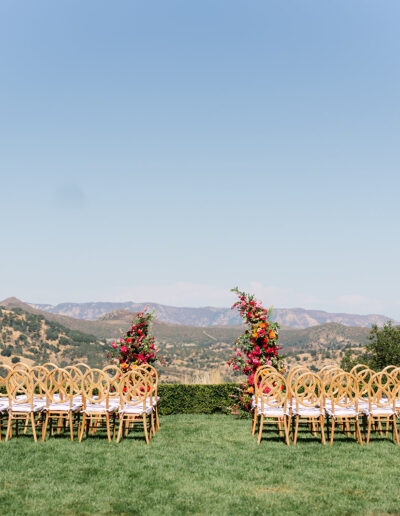 Rows of wooden chairs arranged outdoors on a lawn, facing a floral arch, with mountains and a clear sky in the background.