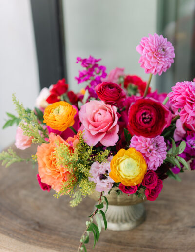A floral arrangement in a silver vase featuring pink roses, yellow, orange, and red ranunculus, dahlias, and green foliage on a wooden table.