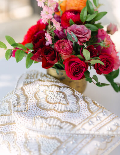A beaded white and gold clutch rests on a table next to a vase of assorted vibrant flowers including roses and greenery.