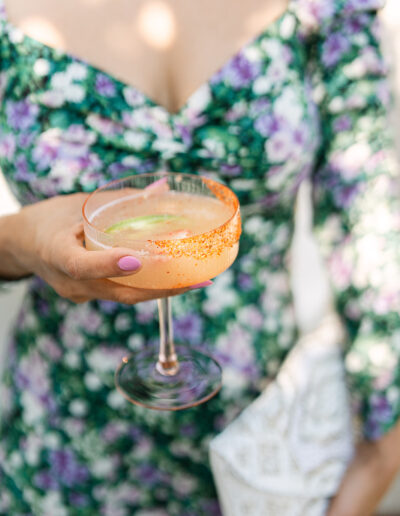 A person in a green and purple floral dress holds a cocktail glass with an orange rim and a light drink, with a white textured clutch in their other hand.