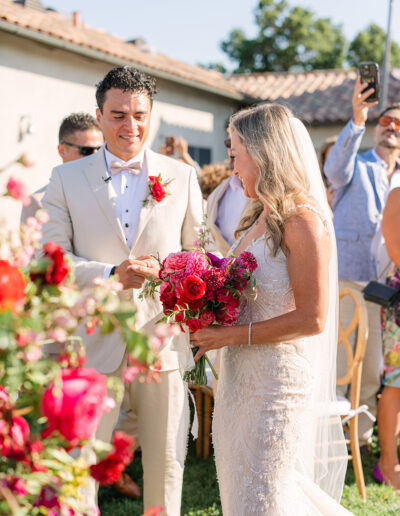 A bride in a lace dress holds a bright bouquet and stands beside a groom in a beige suit during an outdoor wedding ceremony.