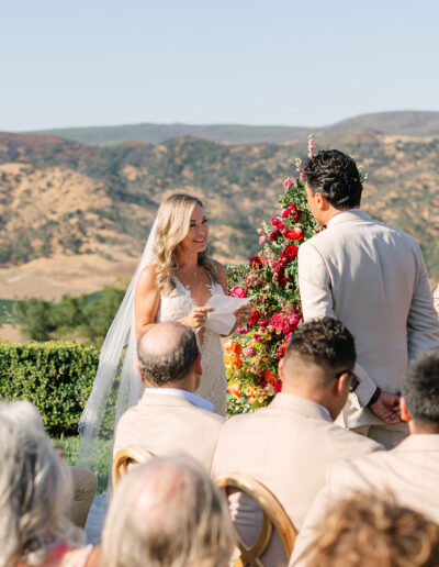 A bride and groom stand facing each other outdoors during their wedding ceremony, with guests seated and a colorful floral arrangement behind them.