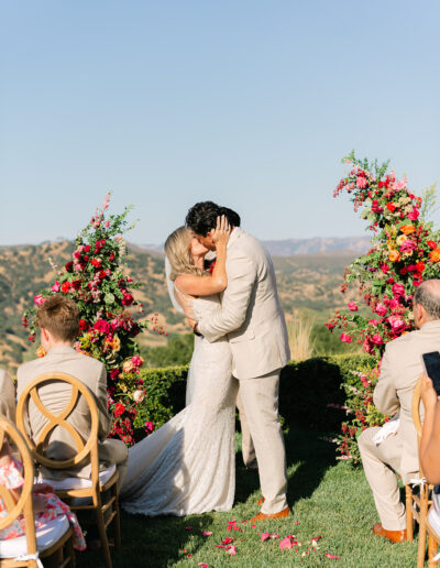 A bride and groom kiss at an outdoor wedding ceremony, surrounded by colorful floral arrangements and seated guests, with mountains in the background.