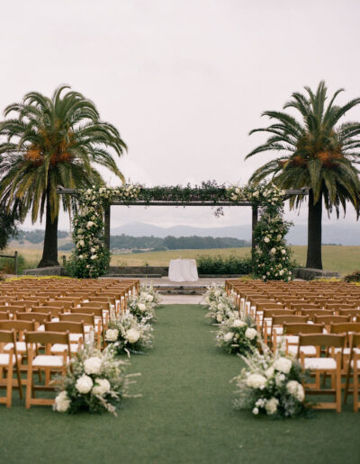 Outdoor wedding venue with rows of wooden chairs, white floral arrangements, and a flower-covered archway framed by two palm trees, set against a scenic landscape.